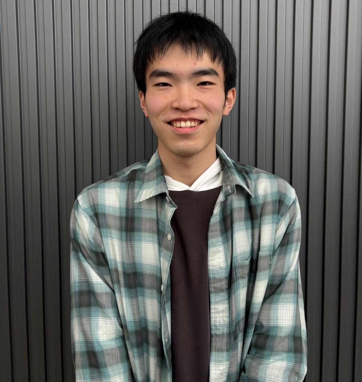 機械システム工学科1年生の平嶋さんが、チェックのシャツを着用し、リラックスした表情でインタビューに応じる様子