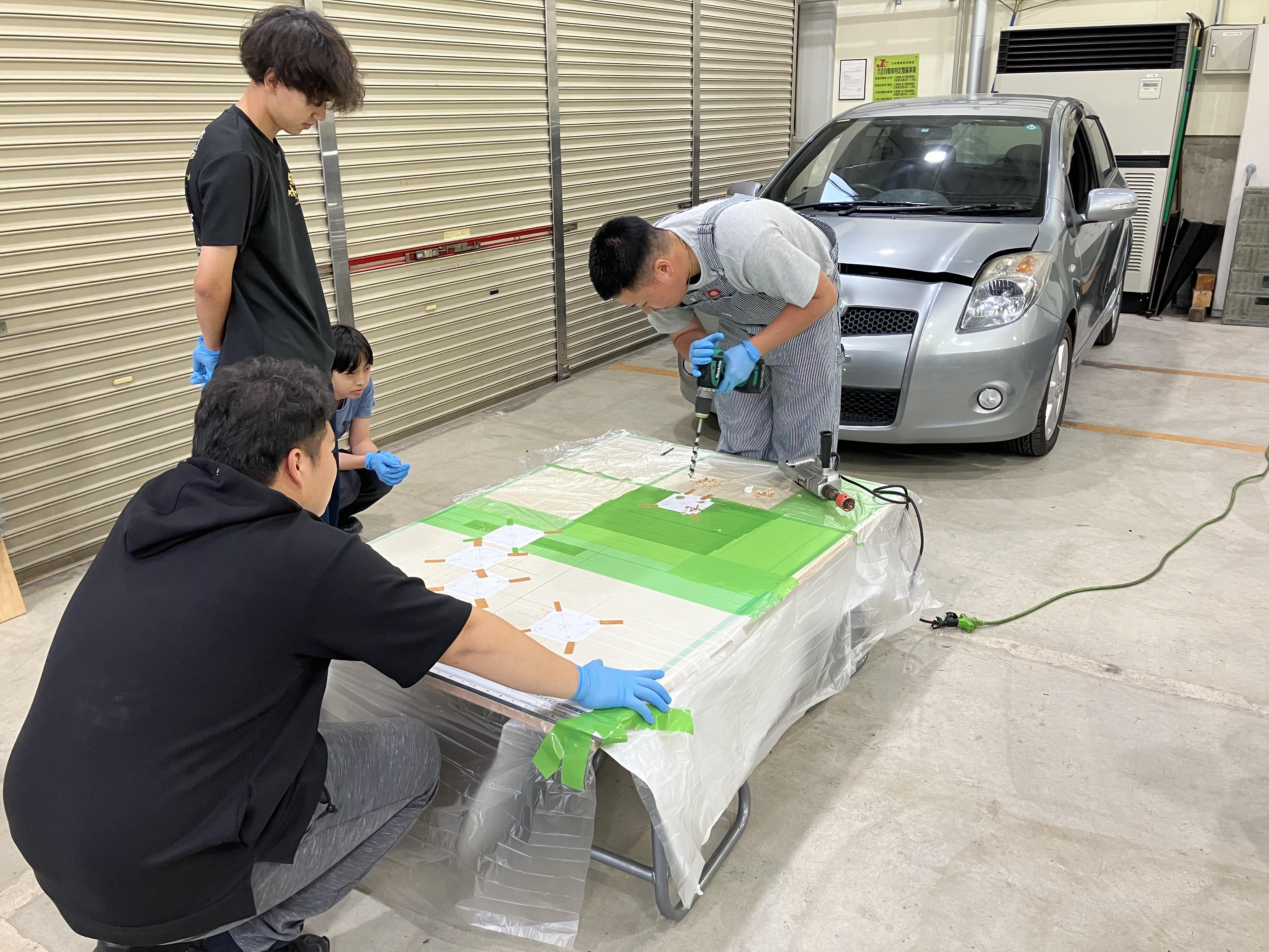 プロジェクトメンバーの学生たちが集まり、作業台の上の展示物を見つめながら、安全性の確認や細かな部品の調整について話し合いながら作業している様子。