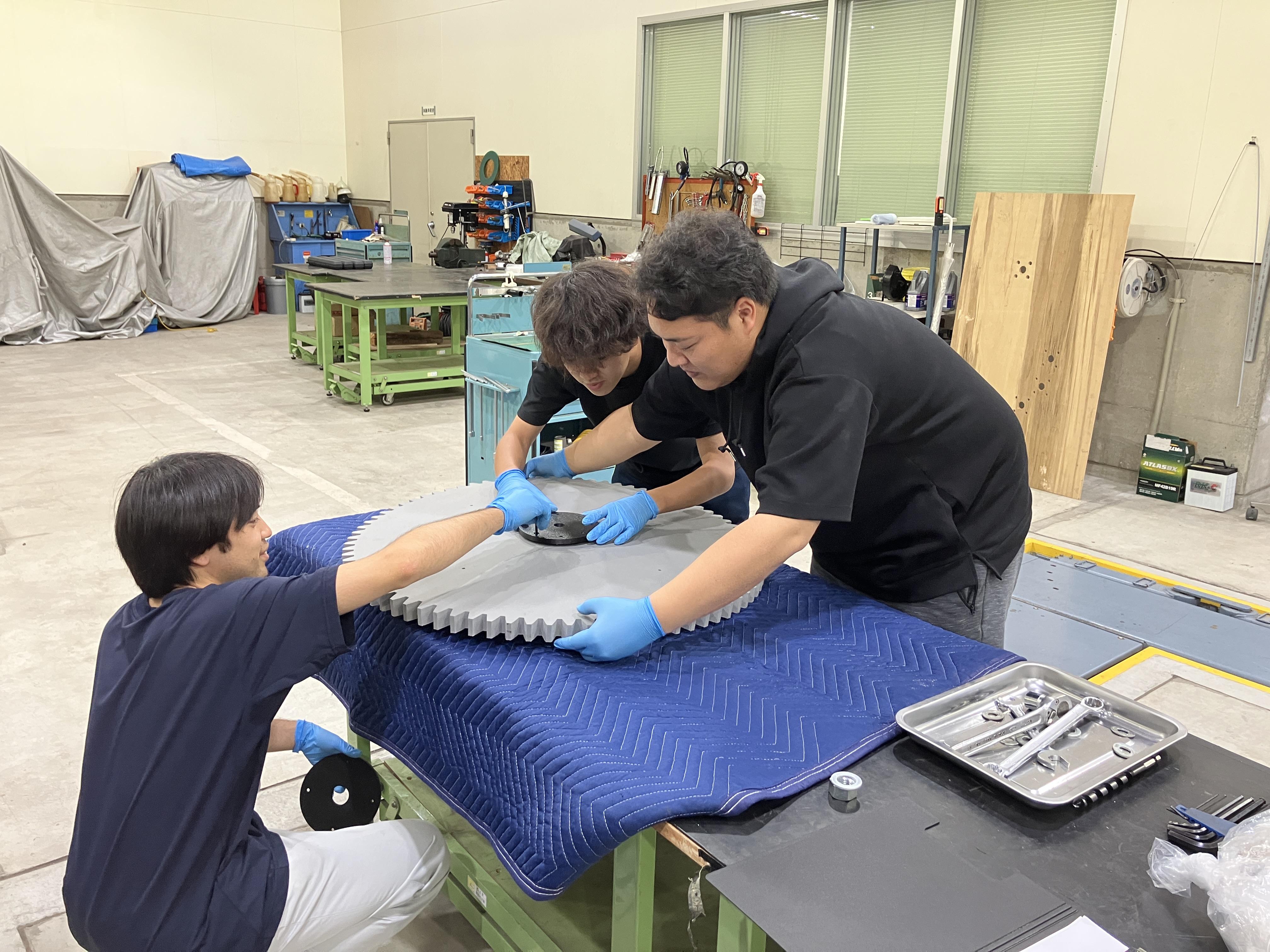 「スケスケ展2」の製作風景。3人のスタッフが大きな歯車を大事そうに抱えながら、協力して慎重に作業を行っている様子。