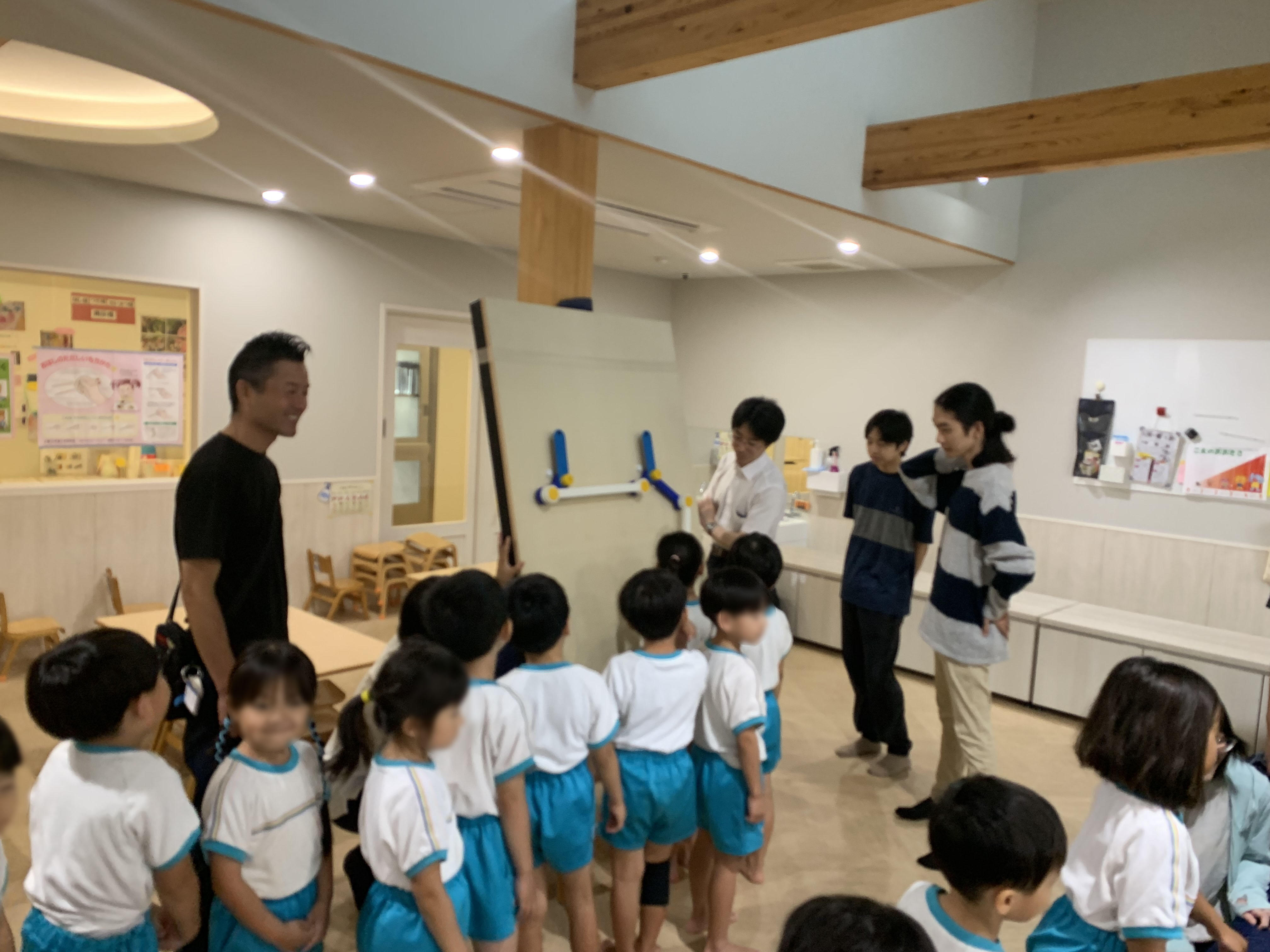 ハンドルを回すと連動して動く、装置の上部(ワイパー構造のパーツ)を園児が指差し、学生が優しく見守っている様子。