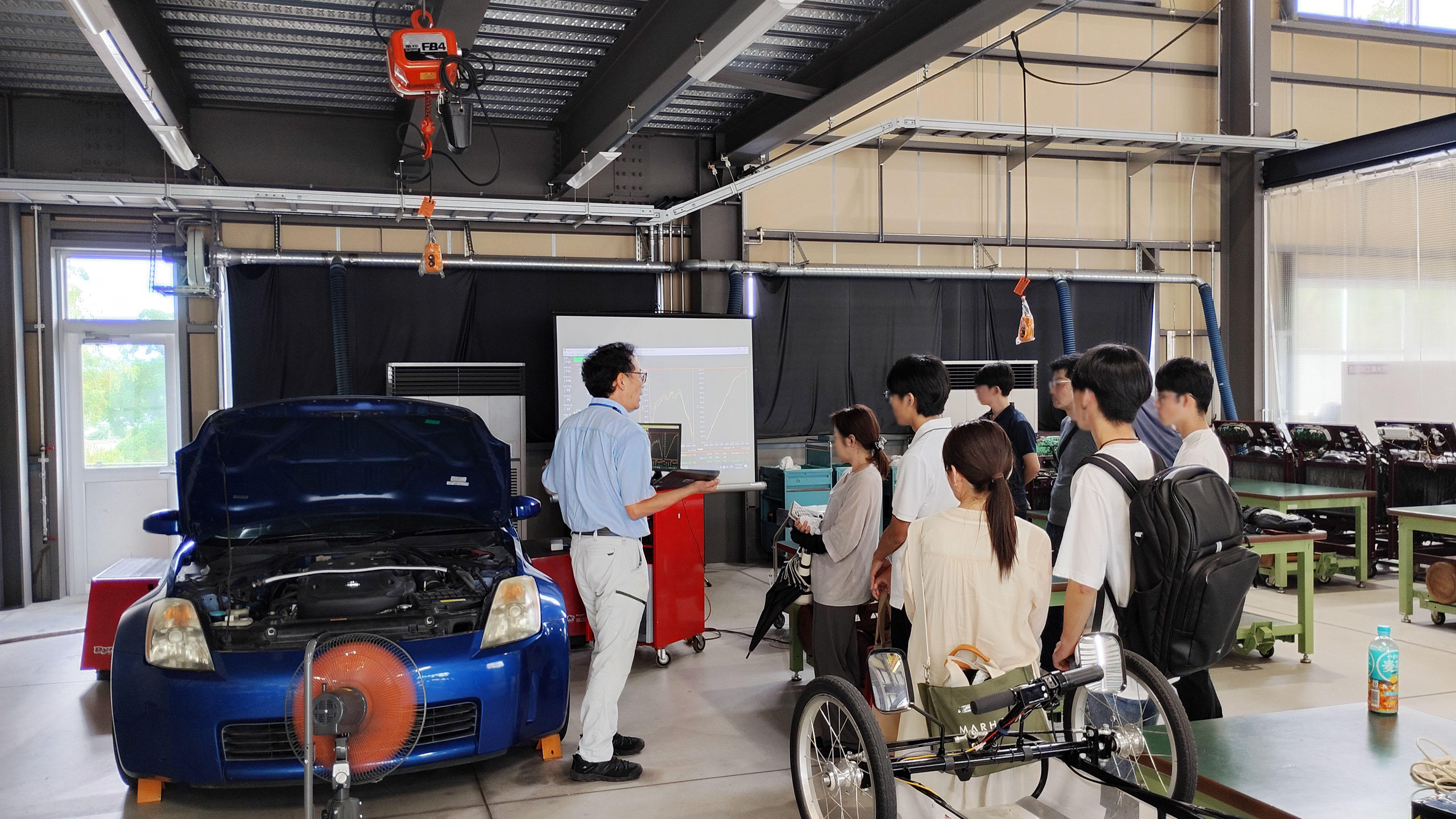 久留米工業大学のオープンキャンパスで、交通機械工学科の体験ブース(スポーツカーの動力性能測定やモビリティ技術)に集まり熱心に解説を聞く参加者の様子