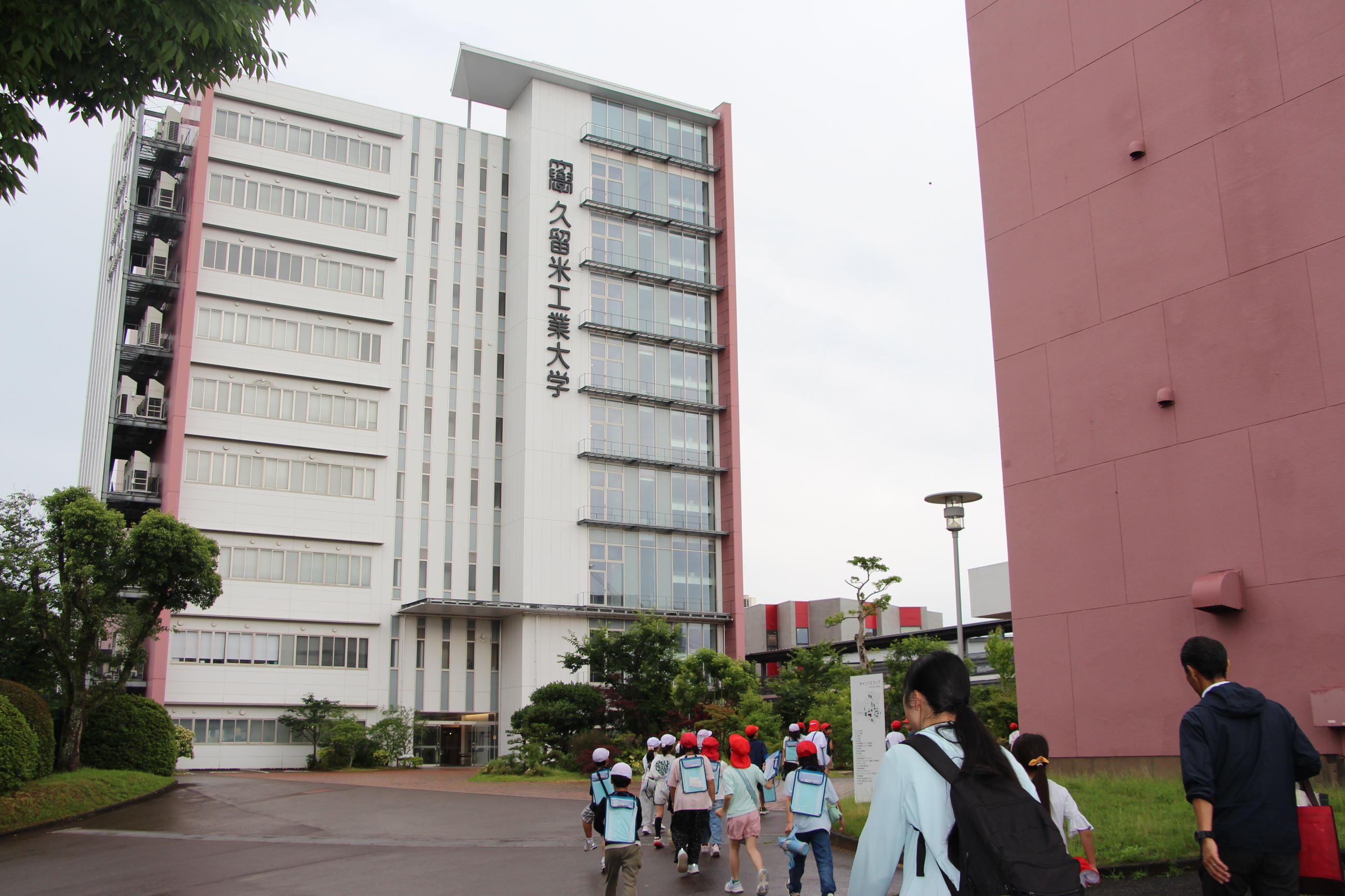 社会科見学で久留米工業大学を訪れた上津小学校の児童たちが、引率の教員と共に大学の学内施設へ向かってキャンパス内を移動している風景。