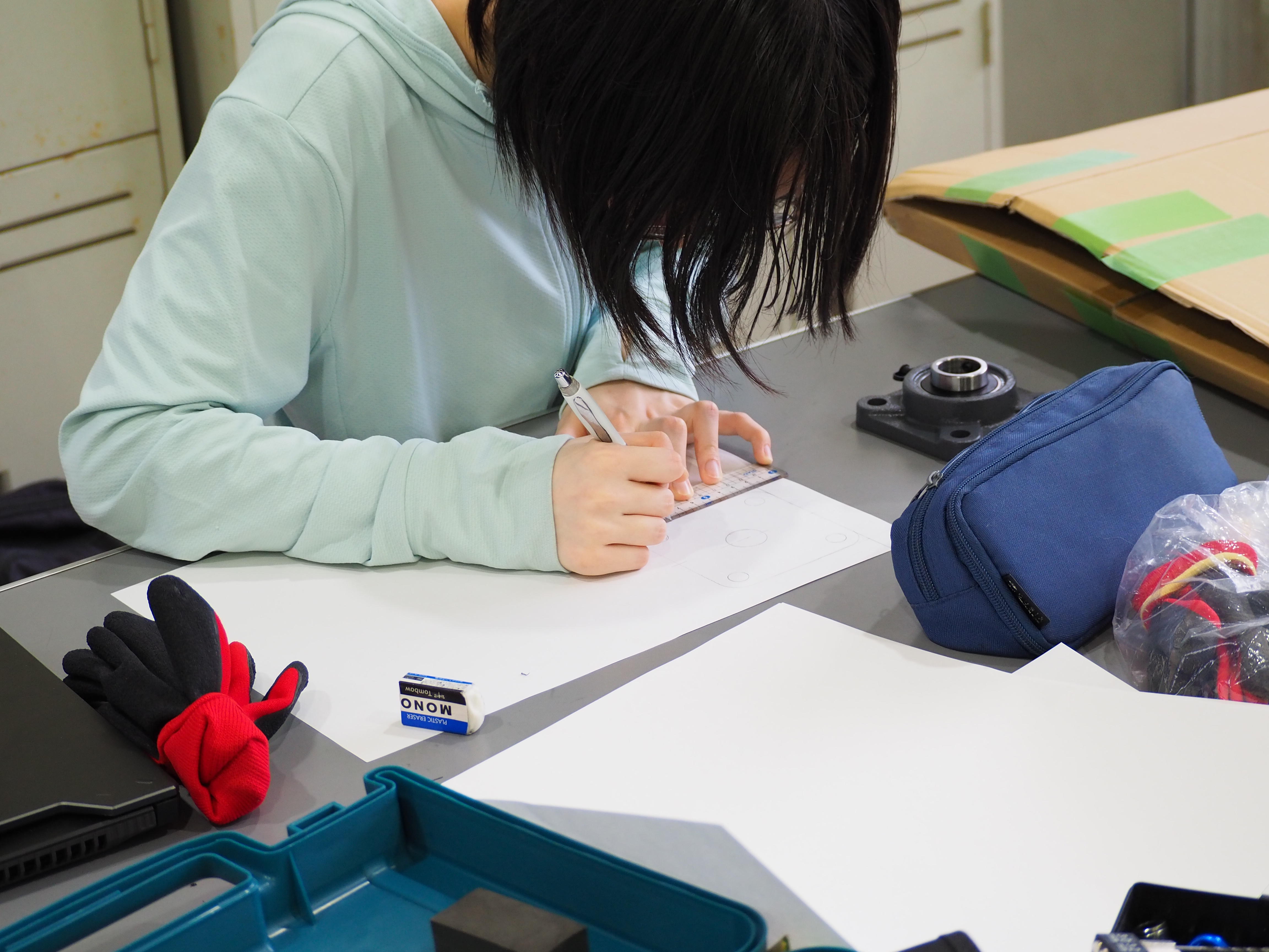 久留米工業大学の学生が机に向かい、紙に文字や図を書きながら展示物のアイデアを練っている作業風景