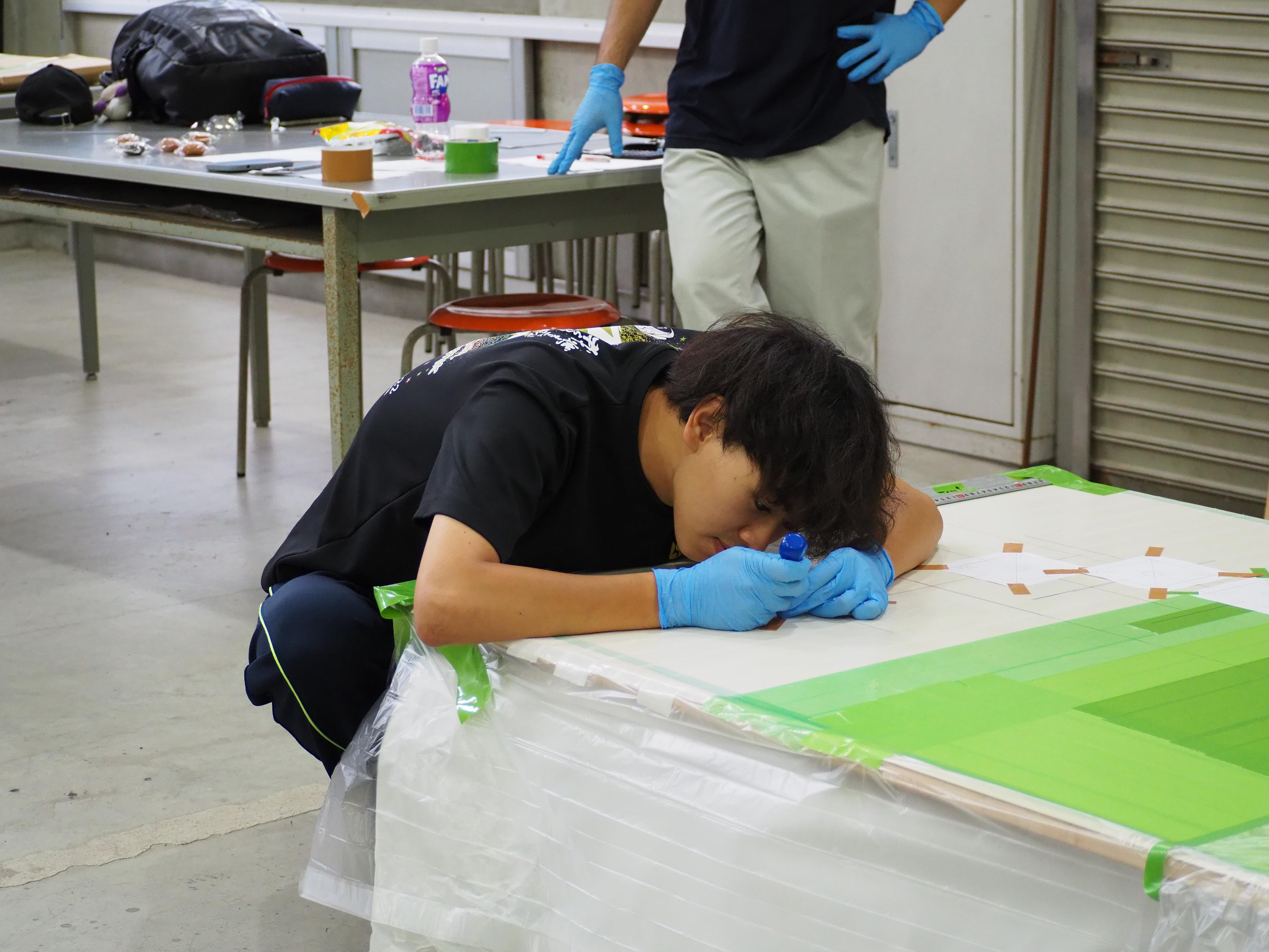 作業台の上に置かれた板状の展示物に対し、かがみ込みながらペン先や工具をあてて、細かな調整や文字の書き込みを丁寧に行っている学生の様子。