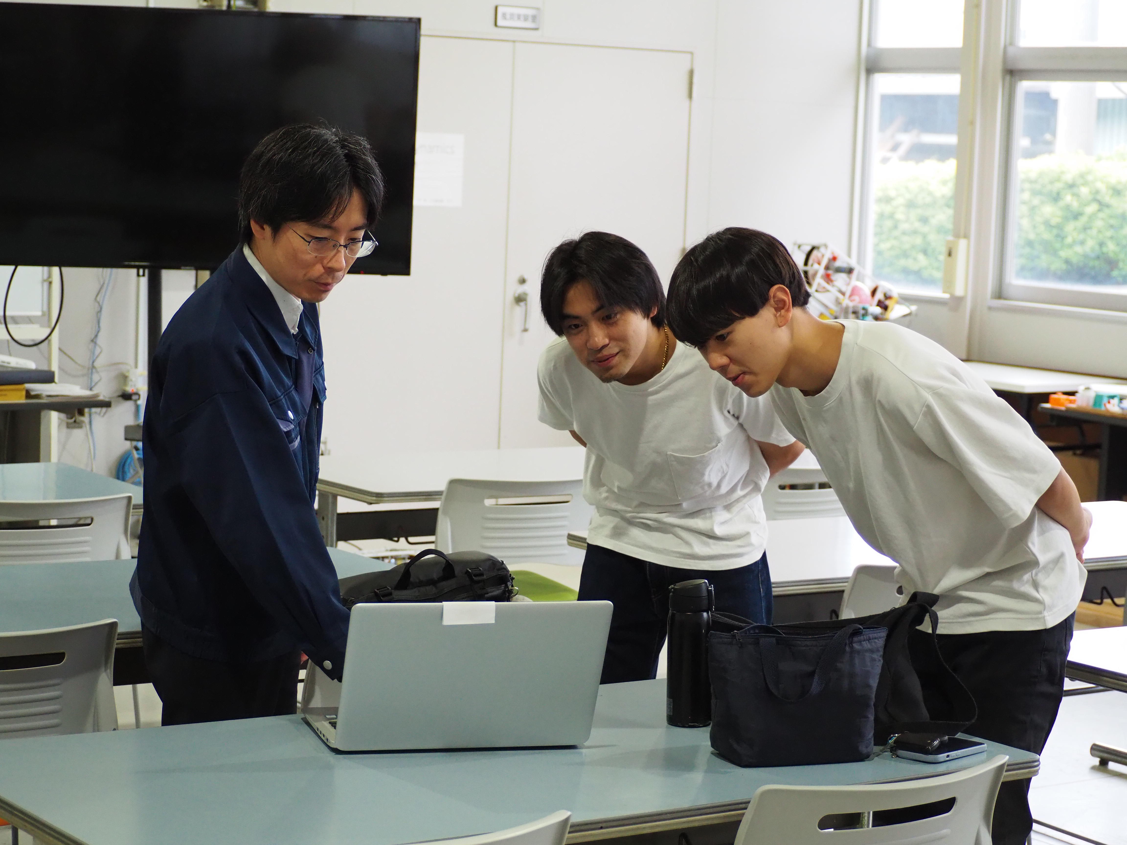 久留米工業大学の教員と学生たちが、パソコンの画面を一緒にのぞき込んで確認を行っている様子