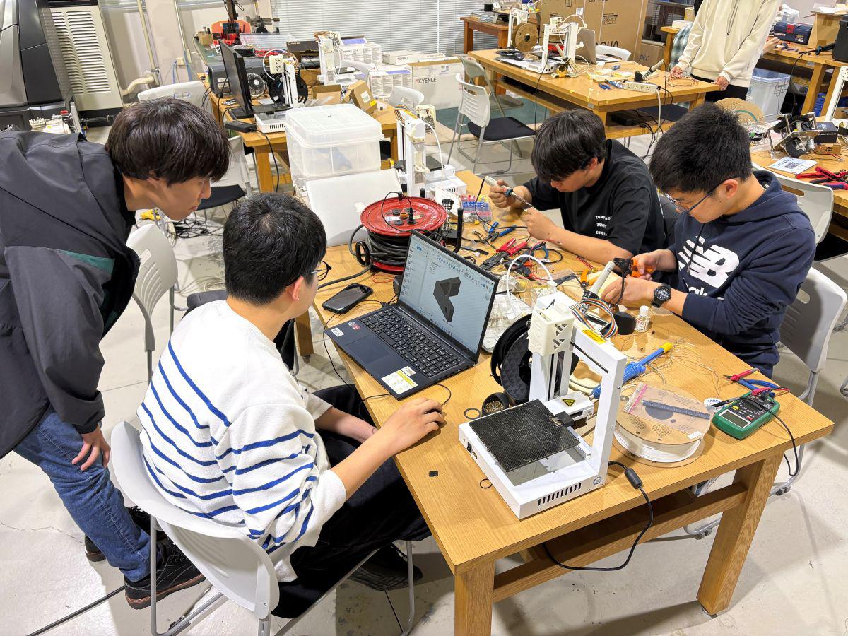 久留米工業大学のものづくりセンターで、パソコンや工具が並ぶ作業台を囲み、相談しながらロボット開発を進める4名の上級生たち
